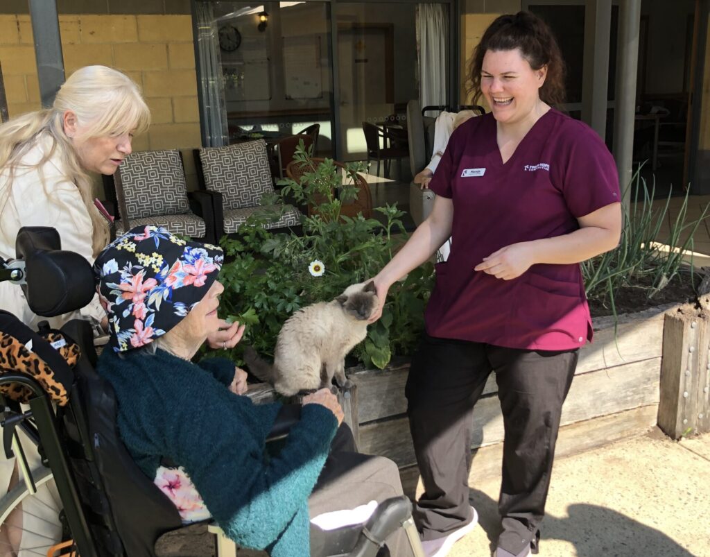 An aged care nurse in a new uniform greets a resident in a wheelchair.